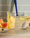 Close up of a GIR Ladle on a white marble surface with a bowl of juice and a glass of juice behind it. 