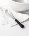 The Black Mini Whisk resting on a white kitchen towel next to a white bowl.