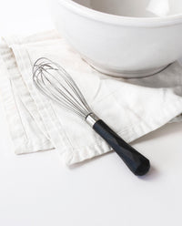 The Black Mini Whisk resting on a white kitchen towel next to a white bowl.