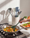 Person cooking vegetables in a pan with a green spatula, kitchen utensils in the background.