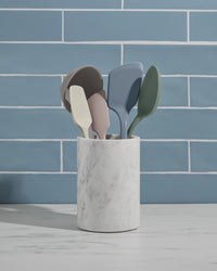 Set of colorful kitchen utensils in a marble holder against a blue tiled wall.