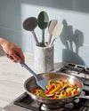 Person using a spatula to stir vegetables in a pan with kitchen utensils in the background.