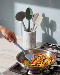 Person using a spatula to stir vegetables in a pan with kitchen utensils in the background.