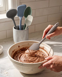 Person mixing chocolate batter in a bowl with a gray spatula, surrounded by colorful spatulas in the background.