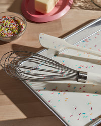 The Sprinkle Baking mat with the Sprinkle Whisk and Spatula on top of it on a wooden surface.. 
