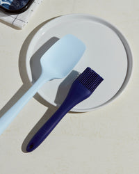The Ultimate Navy Basting Brush next to the Light Blue Spoonula resting on a white plate. 