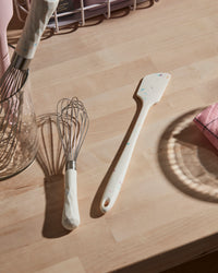 The Sprinkle Skinny Spatula next to the Studio Whisk on a wooden surface. 