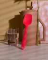 The Red Ultimate Spoonula resting upright on a Matte Sand Ceramic French Press on a creamish background. 