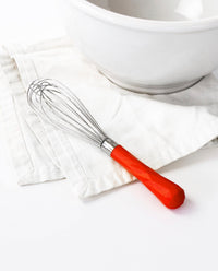 The Red Mini Whisk resting on a white kitchen towel next to a white bowl.