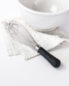 The Black Ultimate Whisk resting on a white kitchen towel next to a white bowl.