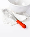 The Red Ultimate Whisk resting on a white kitchen towel next to a white bowl.
