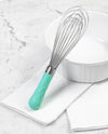 The Sea Foam Ultimate Whisk resting on a white kitchen towel next to a white bowl.