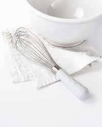 The Studio Ultimate Whisk resting on a white kitchen towel next to a white bowl.