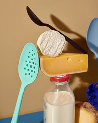 The Sea Foam Perforated Spoon and Mini Black Spoon placed on top on a bottle of milk and blocks of cheese. 