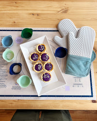 Top view of the Slate Oven Mitts on the GIR Baking mat, next to some cupcakes and Frosty Mint Cupcake Liners on a wooden surface. 